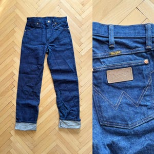 Op de afbeelding: Donkerblauwe denim jeans met een rechte pijp en een knoopsluiting. De jeans hebben een klassiek vijf-zakken ontwerp en een leren patch op de achterzak met het "Wrangler" logo. De manchetten zijn opgerold.
