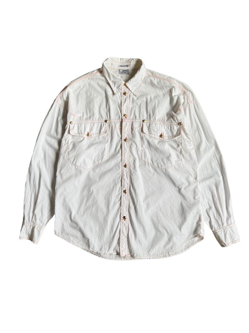 May include: A long-sleeve button-down shirt in off-white fabric with contrasting orange stitching. The shirt features two chest pockets with gold-toned metal buttons, a standard collar, and a Gianni Versace label at the inner neckline.