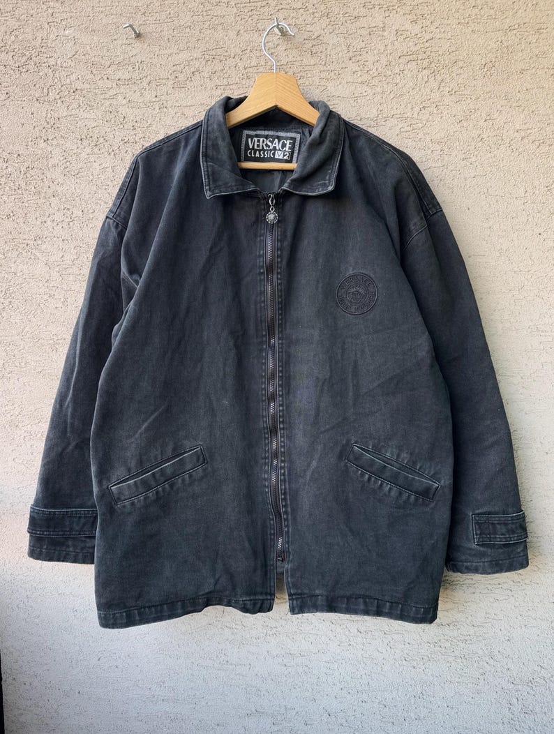 Puede incluir: Chaqueta vaquera gris oscuro con cremallera completa, dos bolsillos delanteros y un peque&ntilde;o parche con el logotipo. La chaqueta tiene cuello y un emblema redondo en el pecho. La etiqueta dice "VERSACE CLASSIC V2".