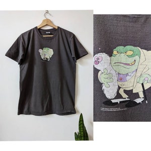 Puede incluir: Una camiseta gris oscuro con una imagen de dibujos animados de una rana verde antropomorfa que lleva un traje y sostiene una criatura blanca y peluda. El texto "© 1991  Copyright  Nelvana Productions Ltd. Licensed by Fremantle Media Licensing. All Rights Reserved." está impreso en la parte inferior de la camiseta.