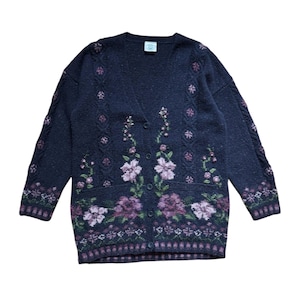 Op de afbeelding: Een donker marineblauwe cardigan met een V-hals en knoopsluiting. De trui heeft een bloemenpatroon in paars, roze en groen. De mouwen hebben een kabelgebreid ontwerp. Een klein label staat bij de halslijn.