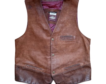 May include: A brown leather vest with a V-neck and four buttons. The vest features two front pockets and a darker brown bottom panel. The interior lining is a deep burgundy colour. The vest has a label that reads "KUDSAK".