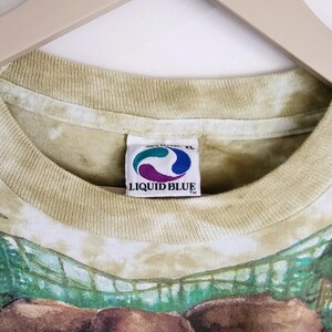Puede incluir: Camiseta Liquid Blue XL con un gr&aacute;fico detallado de elefante. La camiseta tiene un efecto tie-dye en tonos verdes y beige. La etiqueta Liquid Blue es visible en el cuello. La camiseta est&aacute; colgada en una percha de madera.
