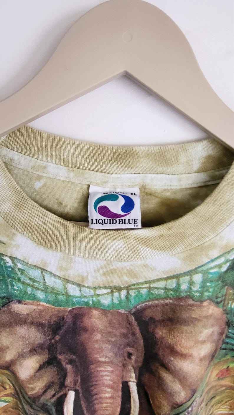 Puede incluir: Camiseta Liquid Blue XL con un gr&aacute;fico detallado de elefante. La camiseta tiene un efecto tie-dye en tonos verdes y beige. La etiqueta Liquid Blue es visible en el cuello. La camiseta est&aacute; colgada en una percha de madera.