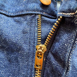 Op de afbeelding: Close-up van een blauwe denim jeans met een messing rits en een knoop. De knoop is lichtbruin van kleur met een logo. De rits is gedeeltelijk open.