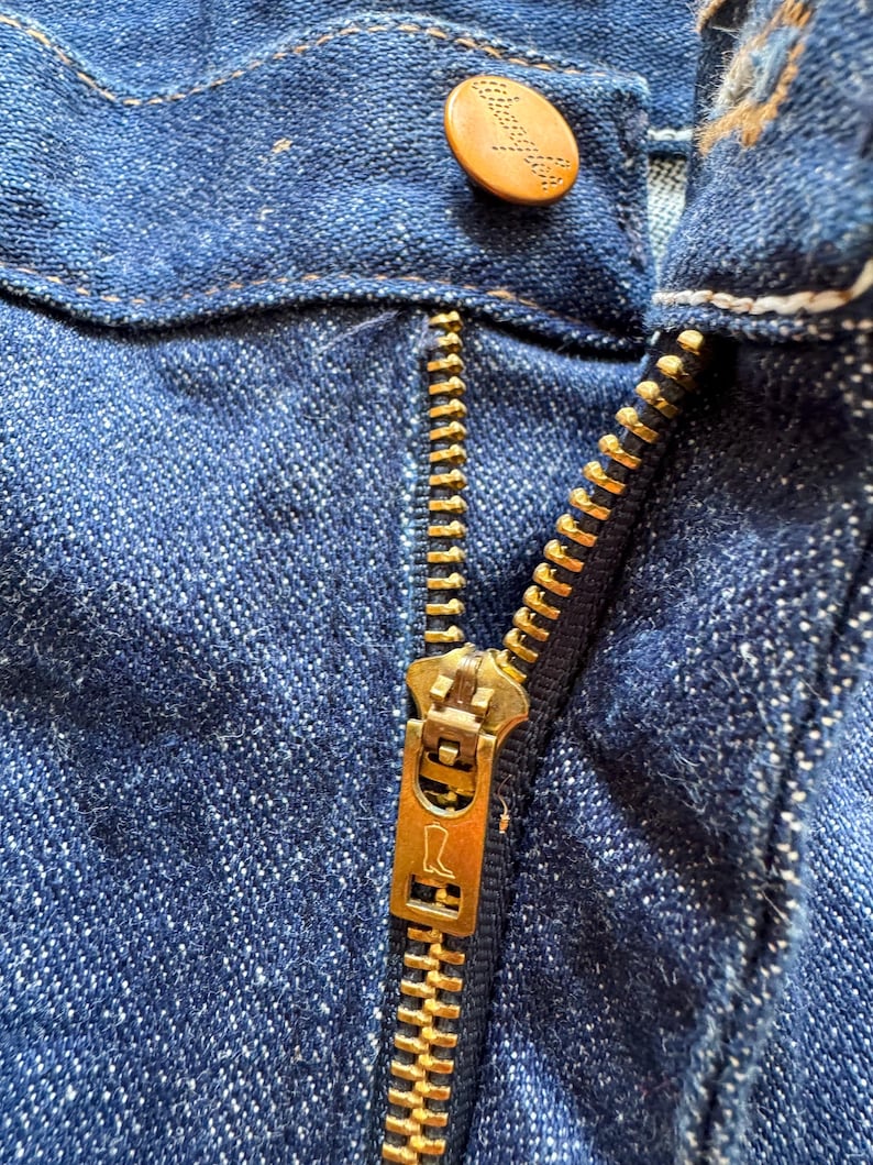 Op de afbeelding: Close-up van een blauwe denim jeans met een messing rits en een knoop. De knoop is lichtbruin van kleur met een logo. De rits is gedeeltelijk open.