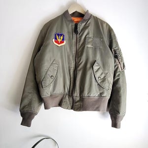 Puede incluir: Chaqueta bomber verde oliva con cierre de cremallera completo, pu&ntilde;os de canal&eacute; y cuello. Cuenta con un parche en el pecho con un emblema alado y un peque&ntilde;o bolsillo en la manga. La chaqueta est&aacute; colgada de una percha de madera.