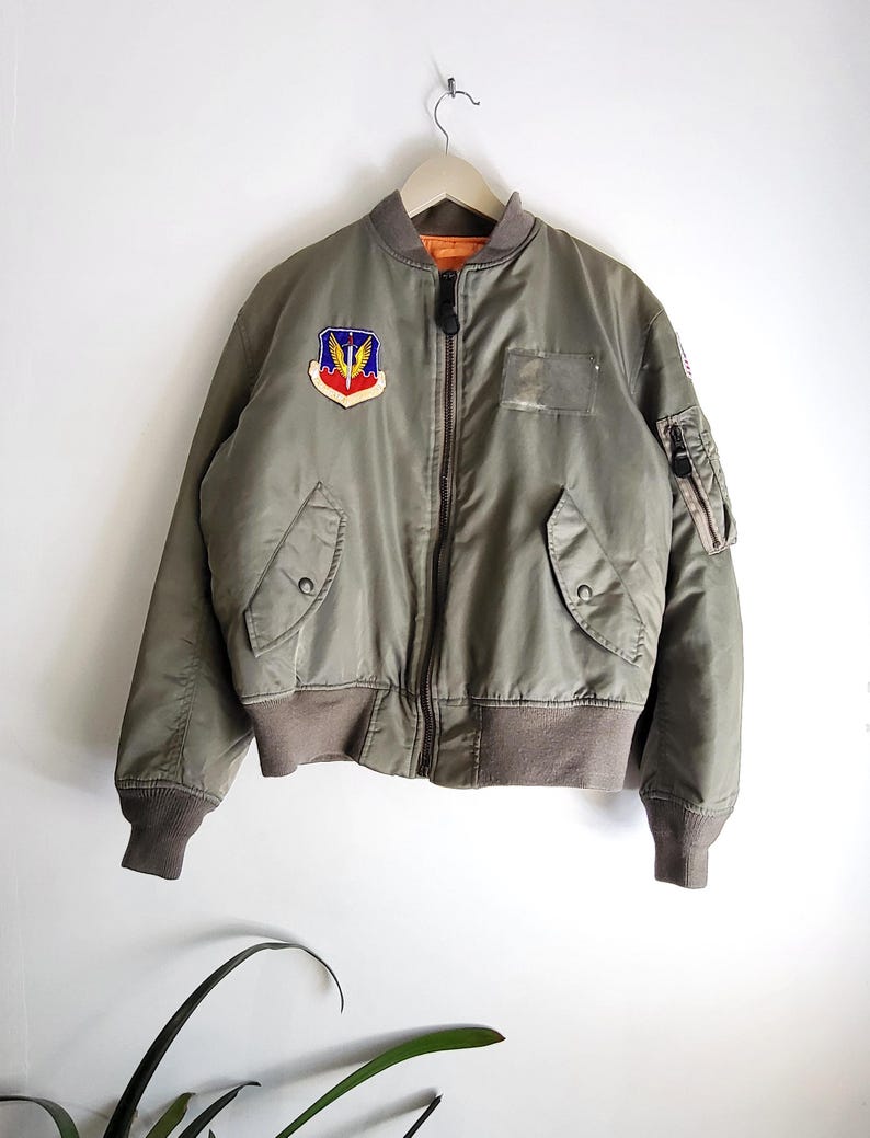 Puede incluir: Chaqueta bomber verde oliva con cierre de cremallera completo, pu&ntilde;os de canal&eacute; y cuello. Cuenta con un parche en el pecho con un emblema alado y un peque&ntilde;o bolsillo en la manga. La chaqueta est&aacute; colgada de una percha de madera.