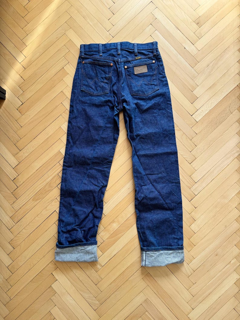 Op de afbeelding: Donkerblauwe spijkerbroek met een klassiek vijf-zakken ontwerp. De jeans heeft een rechte pijp, opgevouwen manchetten en een bruine leren patch op de rug. De stiksels zijn in contrasterend goud.