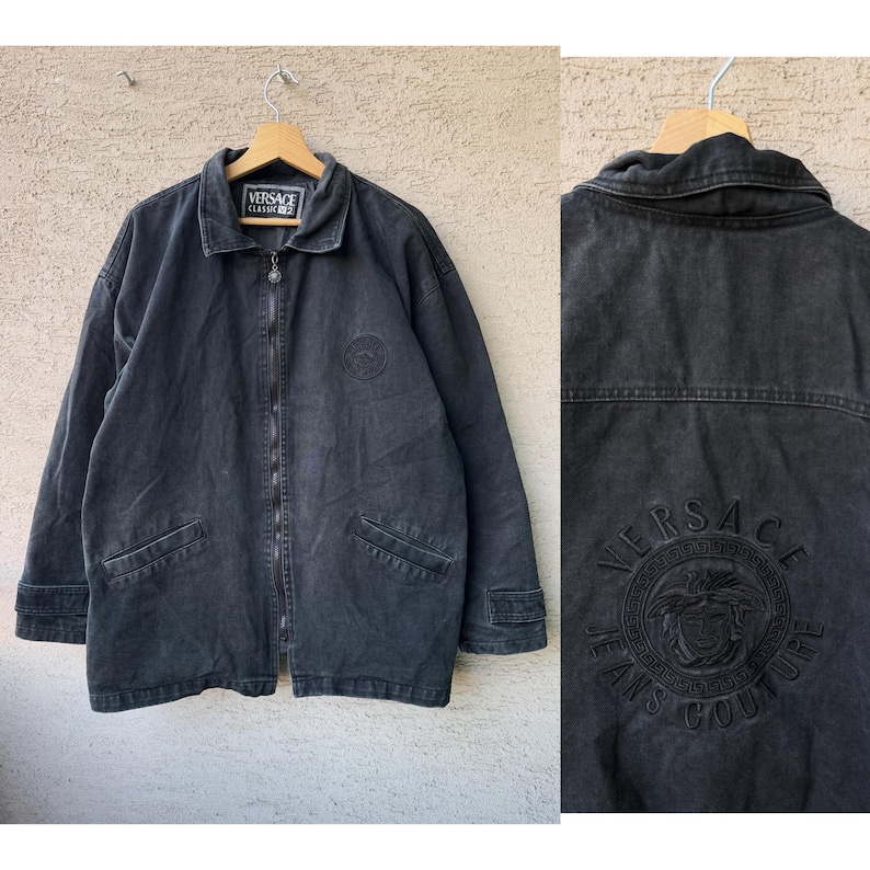 Puede incluir: Chaqueta vaquera negra con cremallera, dos bolsillos delanteros y cuello. La chaqueta presenta el logo de Versace en el pecho y la espalda. La chaqueta est&aacute; colgada de una percha de madera.