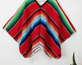 May include: A red, green, blue, and white striped poncho with white fringe. The poncho is hanging on a wooden hanger against a white wall.