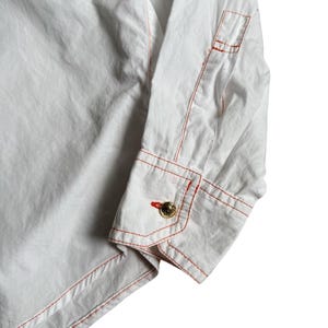 May include: A white long-sleeve shirt featuring prominent orange contrast stitching along the seams and cuffs. The sleeve ends in a structured cuff secured by a single round gold-toned button with an embossed design, complemented by orange buttonhole stitching.