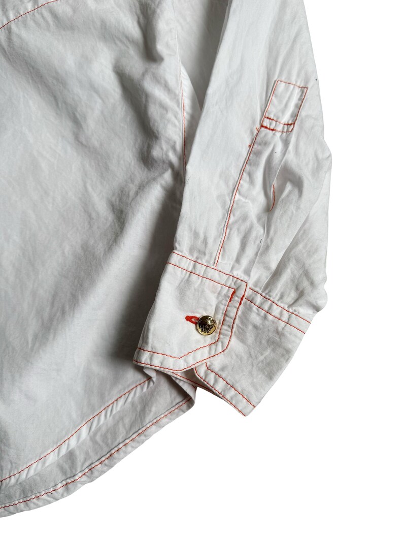 May include: A white long-sleeve shirt featuring prominent orange contrast stitching along the seams and cuffs. The sleeve ends in a structured cuff secured by a single round gold-toned button with an embossed design, complemented by orange buttonhole stitching.