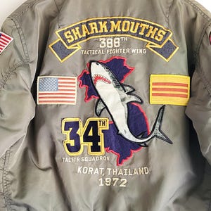 Puede incluir: Chaqueta bomber verde oliva con parches bordados. Presenta un gr&aacute;fico de tibur&oacute;n, el texto "SHARK MOUTHS", "34" y "KORAT, THAILAND 1972". Incluye banderas americanas y otras. La chaqueta tiene cuello y pu&ntilde;os grises.