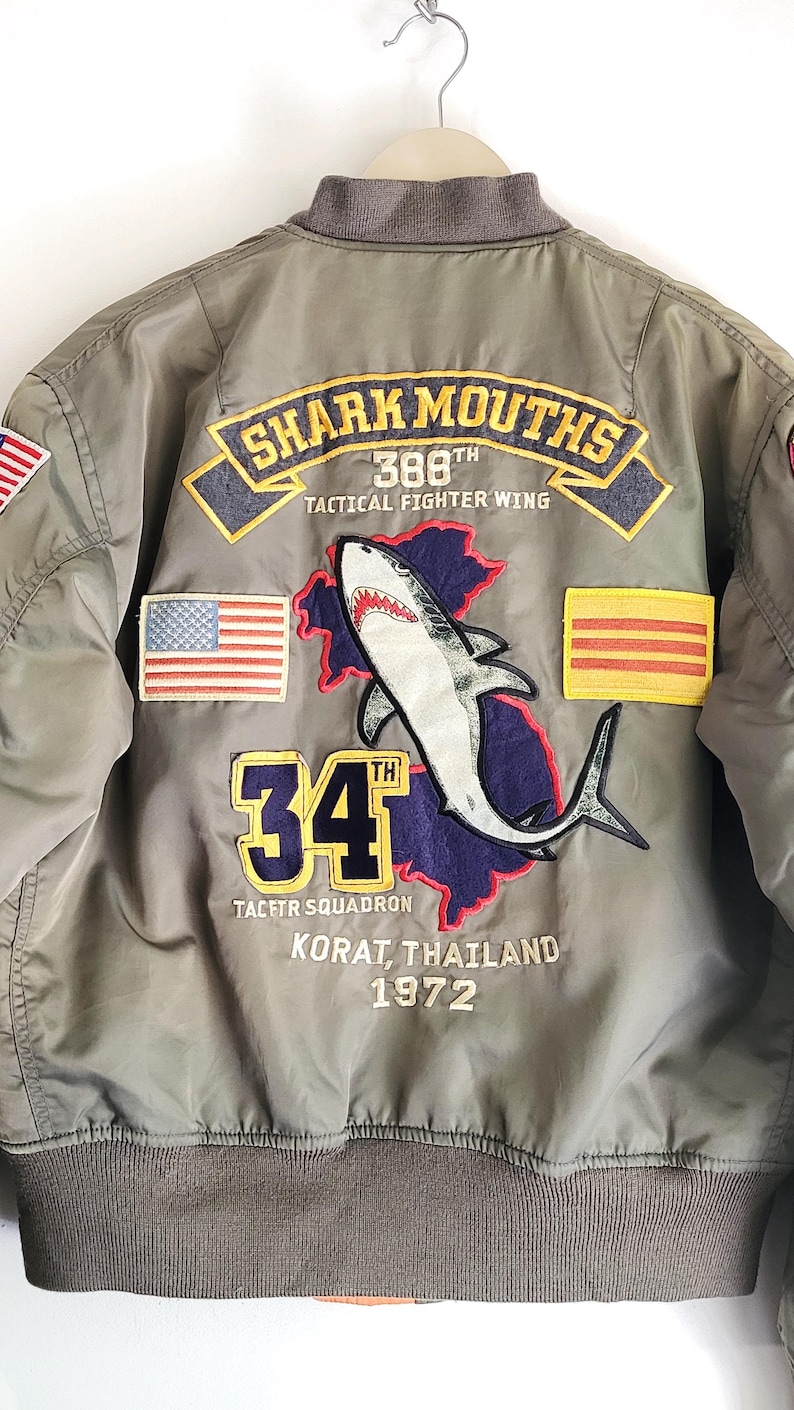 Puede incluir: Chaqueta bomber verde oliva con parches bordados. Presenta un gr&aacute;fico de tibur&oacute;n, el texto "SHARK MOUTHS", "34" y "KORAT, THAILAND 1972". Incluye banderas americanas y otras. La chaqueta tiene cuello y pu&ntilde;os grises.