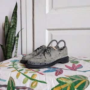 Peut inclure: Une paire de chaussures en cuir gris avec un motif peau de serpent. Les chaussures ont des lacets noirs et une semelle noire. Les chaussures sont posées sur un tissu blanc à motif floral.