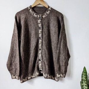 May include: A brown cardigan with a white patterned design around the neckline, cuffs, and hem. The cardigan has a button closure.