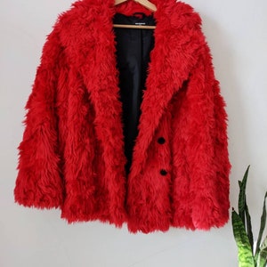 Puede incluir: Una chaqueta de piel sint&eacute;tica roja con cierre de bot&oacute;n negro. La chaqueta est&aacute; colgada en una percha de madera.