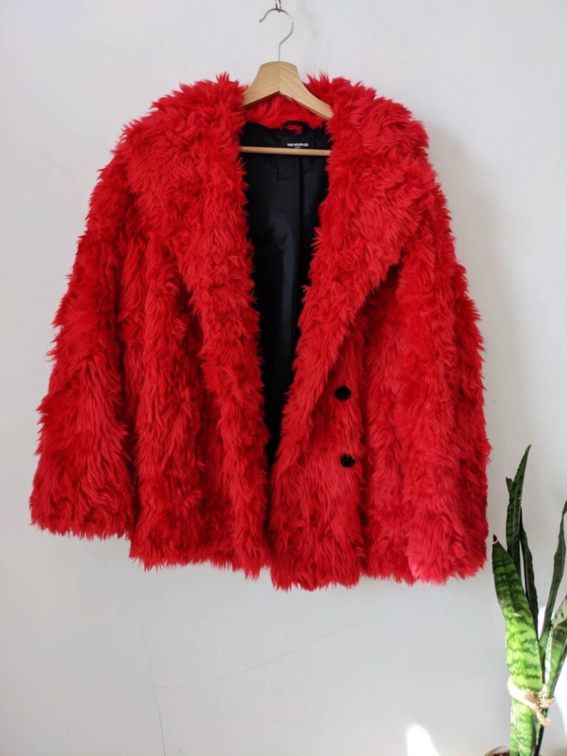 Puede incluir: Una chaqueta de piel sint&eacute;tica roja con cierre de bot&oacute;n negro. La chaqueta est&aacute; colgada en una percha de madera.