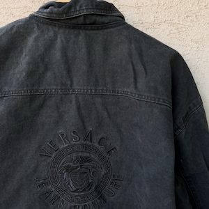 Puede incluir: Chaqueta vaquera negra con el logotipo de Versace Jeans Couture bordado en la espalda. El logotipo presenta el nombre de la marca y un emblema de cabeza de Medusa. La chaqueta est&aacute; colgada en una percha de madera.
