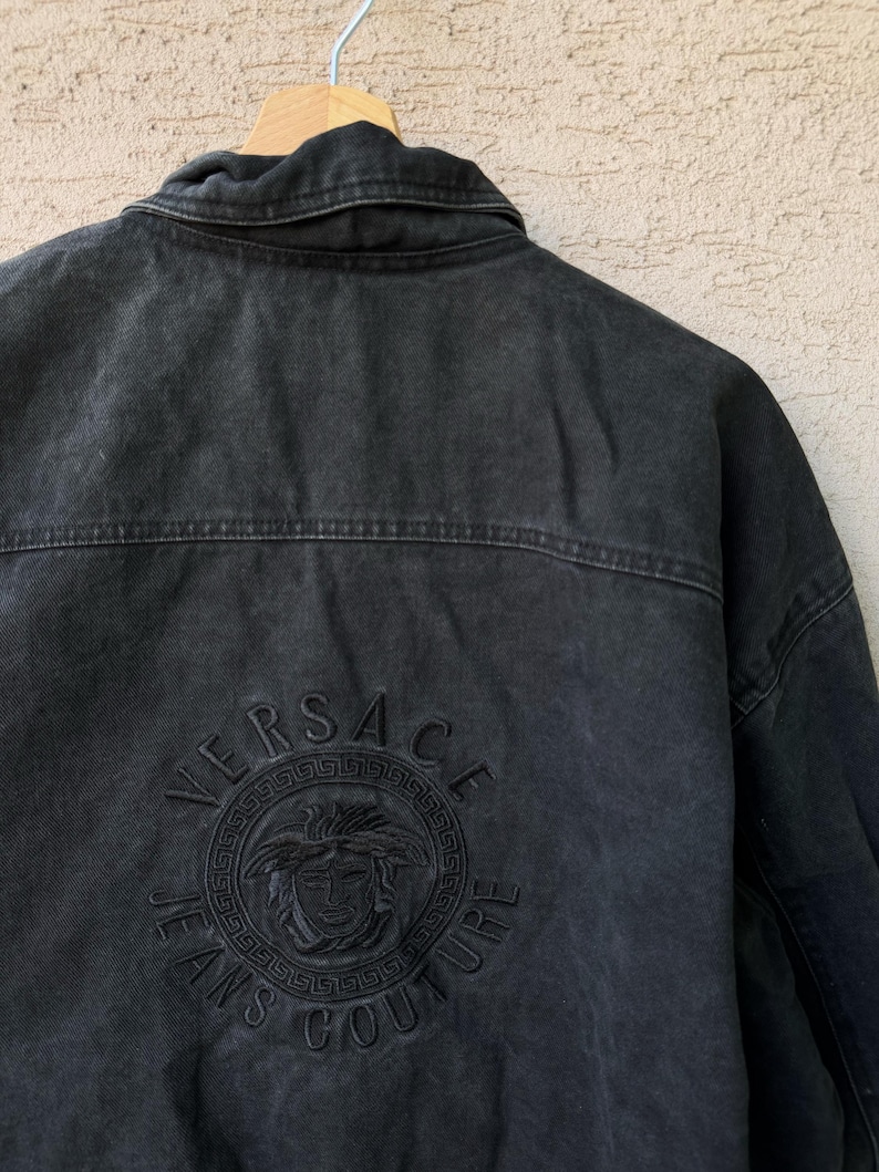 Puede incluir: Chaqueta vaquera negra con el logotipo de Versace Jeans Couture bordado en la espalda. El logotipo presenta el nombre de la marca y un emblema de cabeza de Medusa. La chaqueta est&aacute; colgada en una percha de madera.