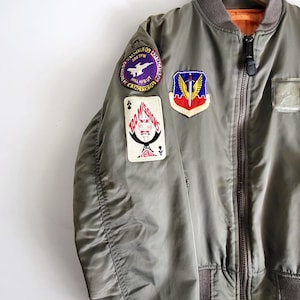Puede incluir: Chaqueta bomber verde oliva con m&uacute;ltiples parches. La chaqueta tiene cuello y pu&ntilde;os marrones, cierre de cremallera e interior naranja claro. Los parches incluyen texto y dise&ntilde;os gr&aacute;ficos.