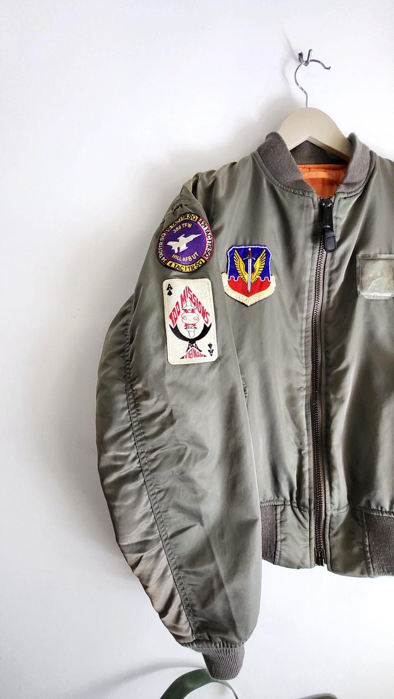 Puede incluir: Chaqueta bomber verde oliva con m&uacute;ltiples parches. La chaqueta tiene cuello y pu&ntilde;os marrones, cierre de cremallera e interior naranja claro. Los parches incluyen texto y dise&ntilde;os gr&aacute;ficos.