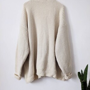 May include: A cream-colored knitted sweater with a ribbed texture. The sweater has long sleeves and a crew neck.