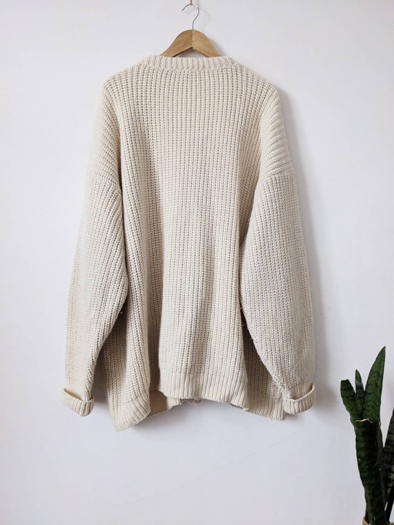 May include: A cream-colored knitted sweater with a ribbed texture. The sweater has long sleeves and a crew neck.