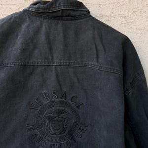 Puede incluir: Chaqueta vaquera negra con el logo de Versace Jeans Couture bordado en la espalda. El logo presenta el nombre Versace, una cabeza de Medusa y las palabras "Jeans Couture" en un dise&ntilde;o circular. La chaqueta est&aacute; colgada en una percha de madera.