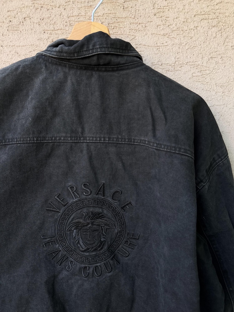 Puede incluir: Chaqueta vaquera negra con el logo de Versace Jeans Couture bordado en la espalda. El logo presenta el nombre Versace, una cabeza de Medusa y las palabras "Jeans Couture" en un dise&ntilde;o circular. La chaqueta est&aacute; colgada en una percha de madera.