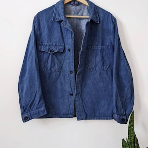 May include: A blue denim work shirt with a button-up front and two large pockets. The shirt is hanging on a wooden hanger against a white wall.