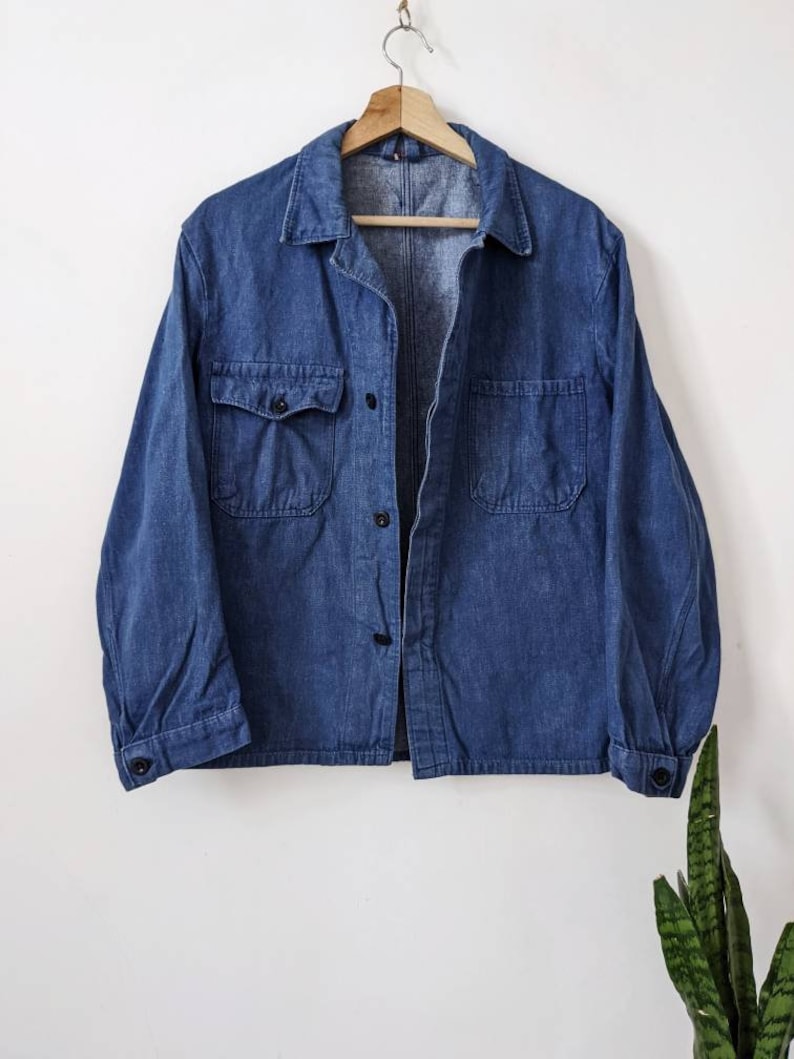 May include: A blue denim work shirt with a button-up front and two large pockets. The shirt is hanging on a wooden hanger against a white wall.