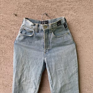 K&ouml;nnte beinhalten: Hellblaue Jeans mit hohem Bund, Knopfverschluss und klassischem F&uuml;nf-Taschen-Design. Die Jeans h&auml;ngen an einem Metallb&uuml;gel und zeigen die Vorderseite des Kleidungsst&uuml;cks. Der Markenname ist am Bund sichtbar.