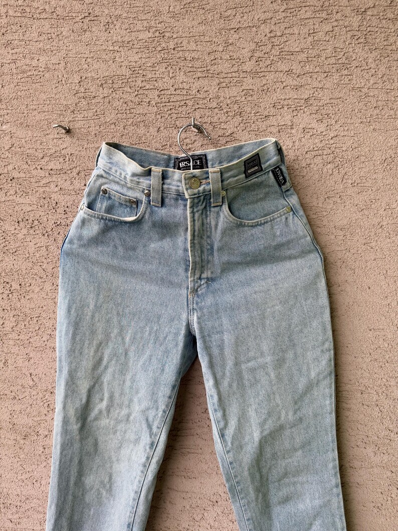 K&ouml;nnte beinhalten: Hellblaue Jeans mit hohem Bund, Knopfverschluss und klassischem F&uuml;nf-Taschen-Design. Die Jeans h&auml;ngen an einem Metallb&uuml;gel und zeigen die Vorderseite des Kleidungsst&uuml;cks. Der Markenname ist am Bund sichtbar.