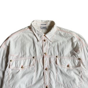 May include: A white long-sleeve button-down shirt featuring contrasting orange stitching throughout. The shirt includes two chest pockets with gold-tone metal buttons, a standard collar, and a label reading GIANNI VERSACE at the neckline.