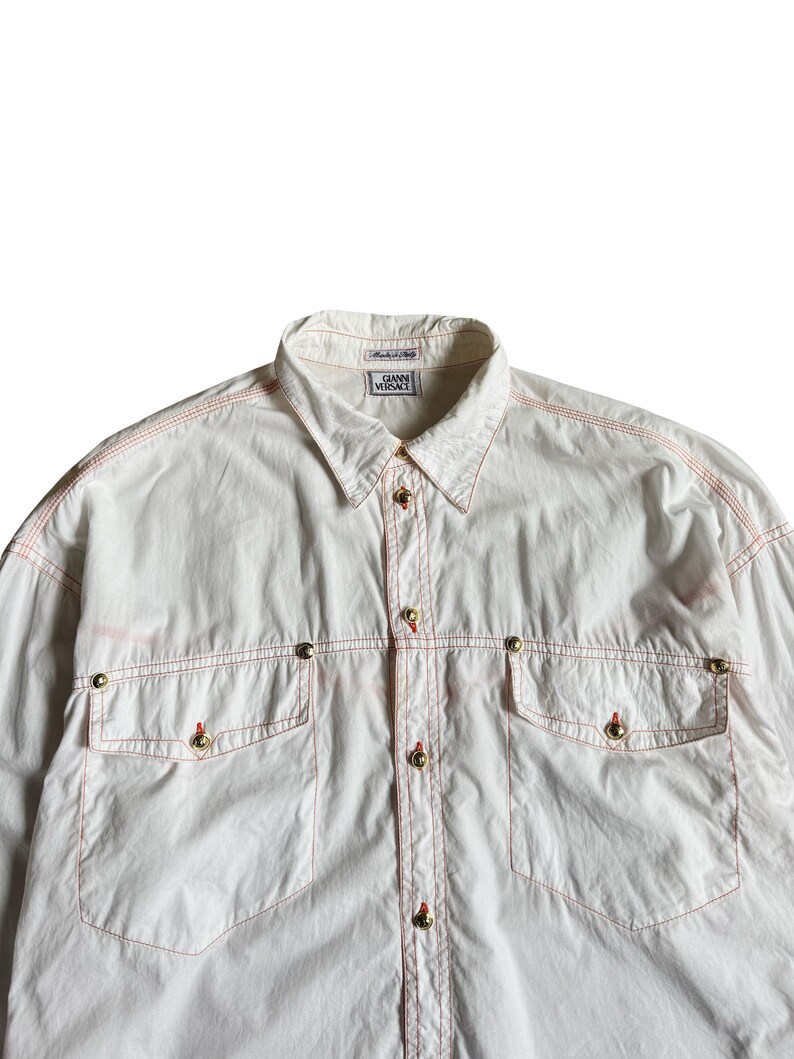May include: A white long-sleeve button-down shirt featuring contrasting orange stitching throughout. The shirt includes two chest pockets with gold-tone metal buttons, a standard collar, and a label reading GIANNI VERSACE at the neckline.
