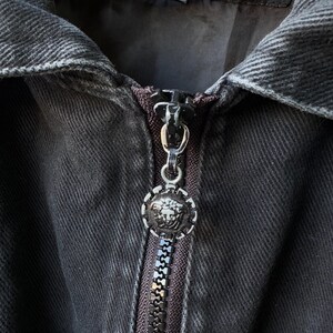 Puede incluir: Primer plano de una chaqueta vaquera negra con cremallera plateada y un tirador redondo decorativo con un emblema de cabeza de Medusa. La chaqueta est&aacute; colgada de una percha de madera, mostrando los detalles de la tela y el dise&ntilde;o.