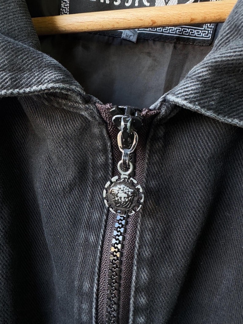 Puede incluir: Primer plano de una chaqueta vaquera negra con cremallera plateada y un tirador redondo decorativo con un emblema de cabeza de Medusa. La chaqueta est&aacute; colgada de una percha de madera, mostrando los detalles de la tela y el dise&ntilde;o.