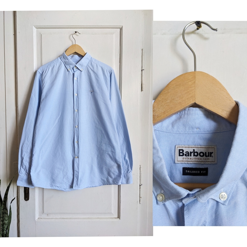 May include: A light blue button-down shirt with long sleeves, hanging on a wooden hanger. The shirt has a small logo on the chest and the brand label "Barbour" is visible. The shirt is tailored fit.