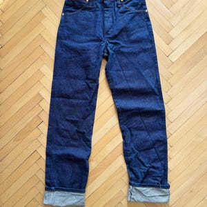 Op de afbeelding: Donkerblauwe denim jeans met een knoopsluiting en goudkleurige klinknagels. De jeans hebben een klassiek ontwerp met vijf zakken en zijn omgeslagen bij de enkels. De broek wordt gepresenteerd op een lichtbruine houten vloer.