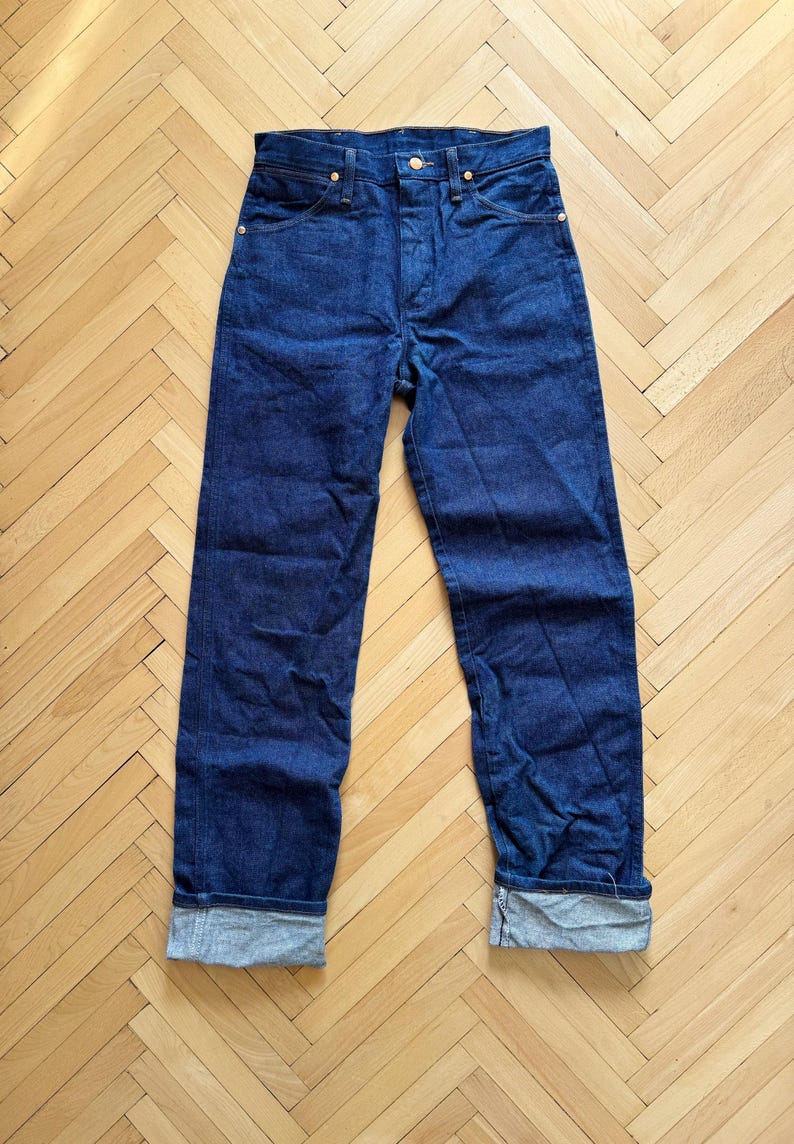 Op de afbeelding: Donkerblauwe denim jeans met een knoopsluiting en goudkleurige klinknagels. De jeans hebben een klassiek ontwerp met vijf zakken en zijn omgeslagen bij de enkels. De broek wordt gepresenteerd op een lichtbruine houten vloer.
