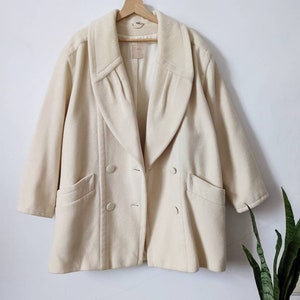 May include: A cream-colored double-breasted wool coat with a large collar and two pockets. The coat is hanging on a wooden hanger against a white wall.