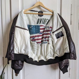 May include: A brown leather jacket with a white interior featuring a map of the United States and the word "AVIREX". The jacket has a black ribbed hem and cuffs, and is hanging on a wooden hanger.