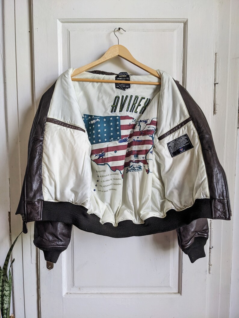 May include: A brown leather jacket with a white interior featuring a map of the United States and the word "AVIREX". The jacket has a black ribbed hem and cuffs, and is hanging on a wooden hanger.