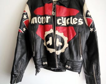 May include: Black leather motorcycle jacket with red and cream-colored accents. The jacket features the words "motor cycles" in a banner across the chest, along with a gear-shaped emblem. The jacket has multiple zippers and pockets.