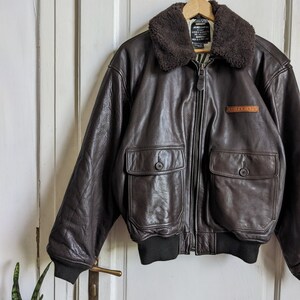 May include: A brown leather bomber jacket with a dark brown shearling collar. The jacket has two front pockets with button closures, a zipper closure, and a black ribbed hem and cuffs. The jacket is hanging on a wooden hanger.