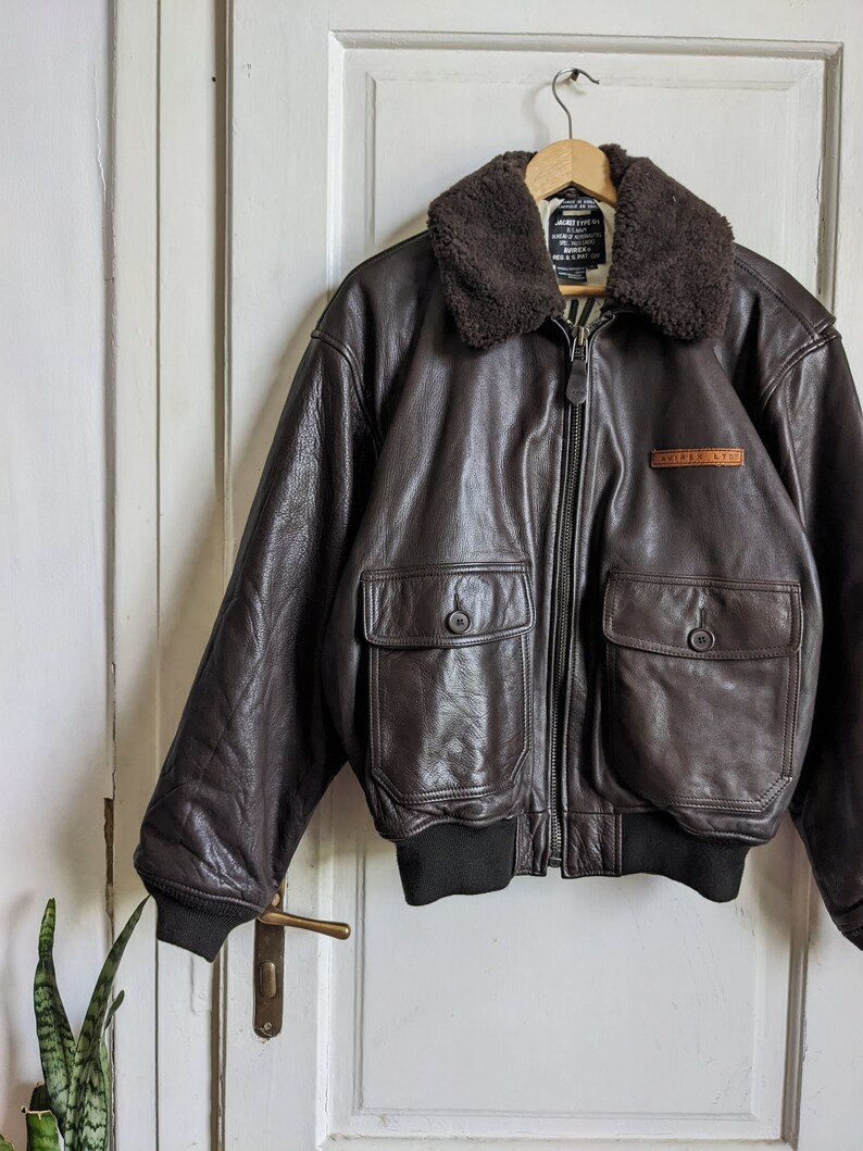 May include: A brown leather bomber jacket with a dark brown shearling collar. The jacket has two front pockets with button closures, a zipper closure, and a black ribbed hem and cuffs. The jacket is hanging on a wooden hanger.