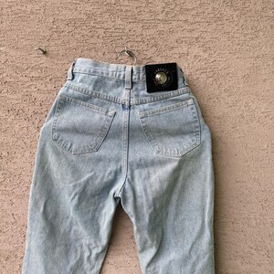 K&ouml;nnte beinhalten: Hellblaue Jeans mit hohem Bund, Ges&auml;&szlig;taschen und einem schwarzen Lederpatch mit goldfarbenem Schriftzug. Die Jeans h&auml;ngen an einem Metallb&uuml;gel vor einem strukturierten Hintergrund.