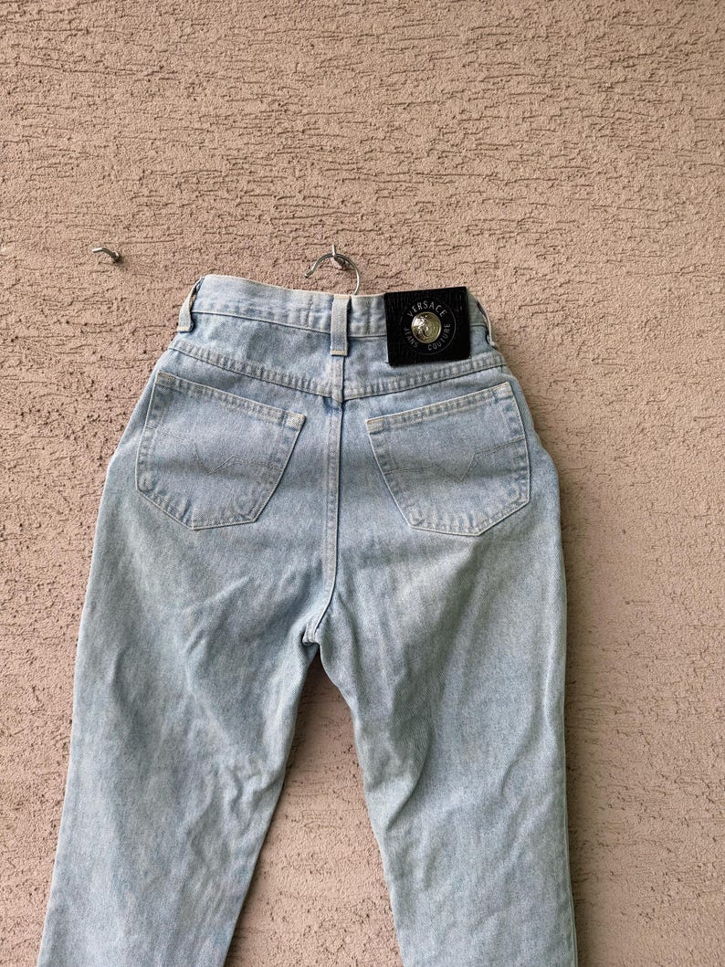 K&ouml;nnte beinhalten: Hellblaue Jeans mit hohem Bund, Ges&auml;&szlig;taschen und einem schwarzen Lederpatch mit goldfarbenem Schriftzug. Die Jeans h&auml;ngen an einem Metallb&uuml;gel vor einem strukturierten Hintergrund.